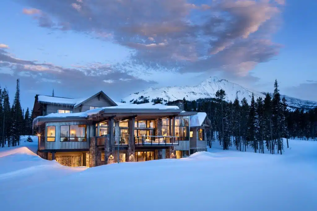 A custom mountain home at dusk, its windows lit beneath a snow-covered peak