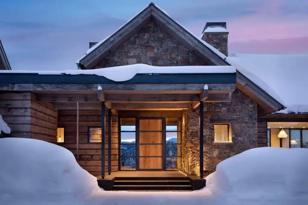 The lit timber-and-stone entry of a snowbound home in Big Sky, Montana, built by Highline Partners